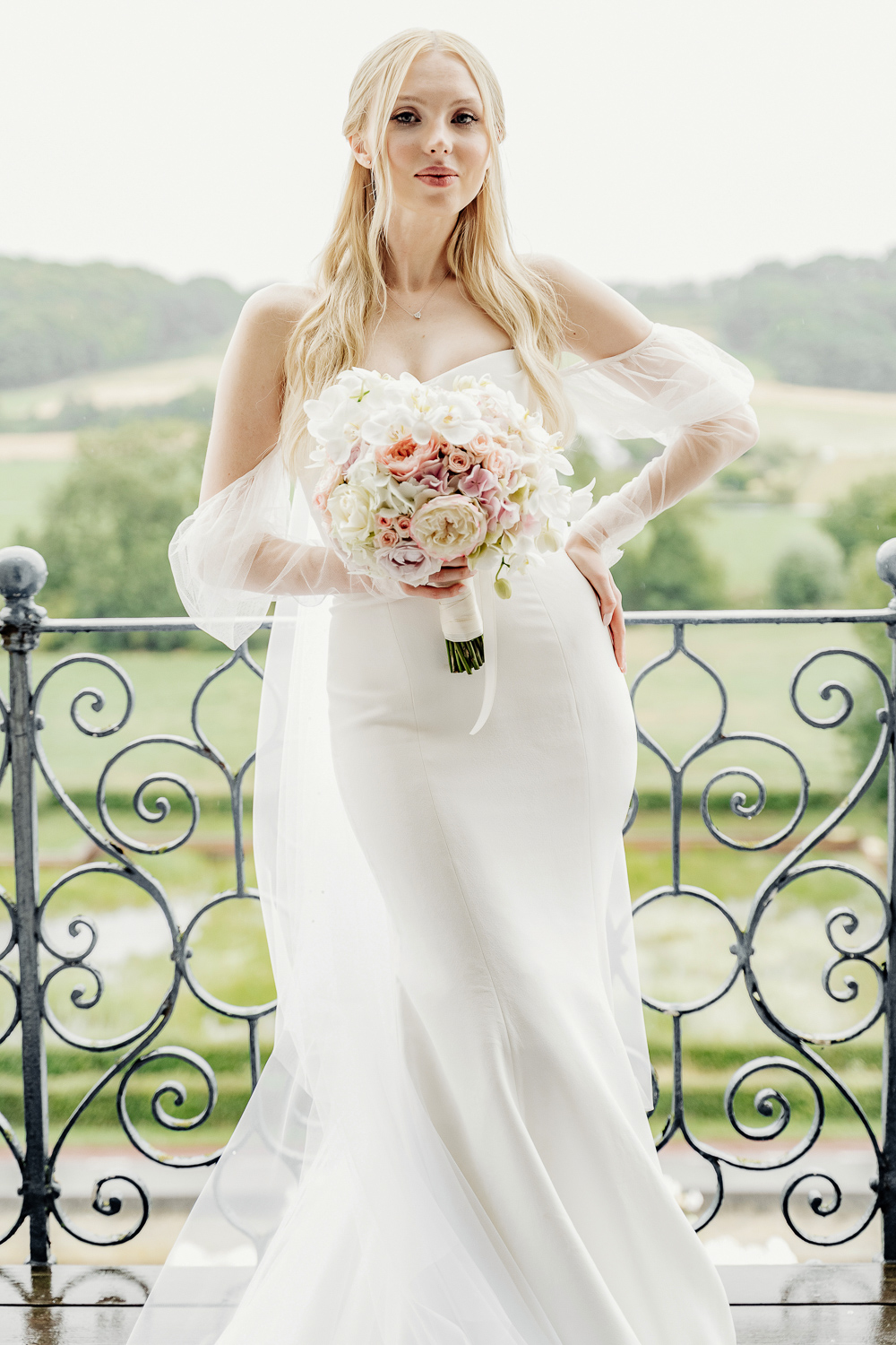 Beautiful blond bride stays in the balcony of castle Chateau Neercanne in Maastricht and slightly smiling, with long veil and wedding dress Vera Wang by Mercury, holding flower bouquet