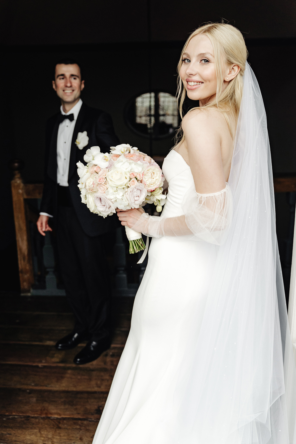 Beautiful blond bride stays in the balcony and slightly smiling and looking back, with long veil and wedding dress Vera Wang by Mercury, holding flower bouquet