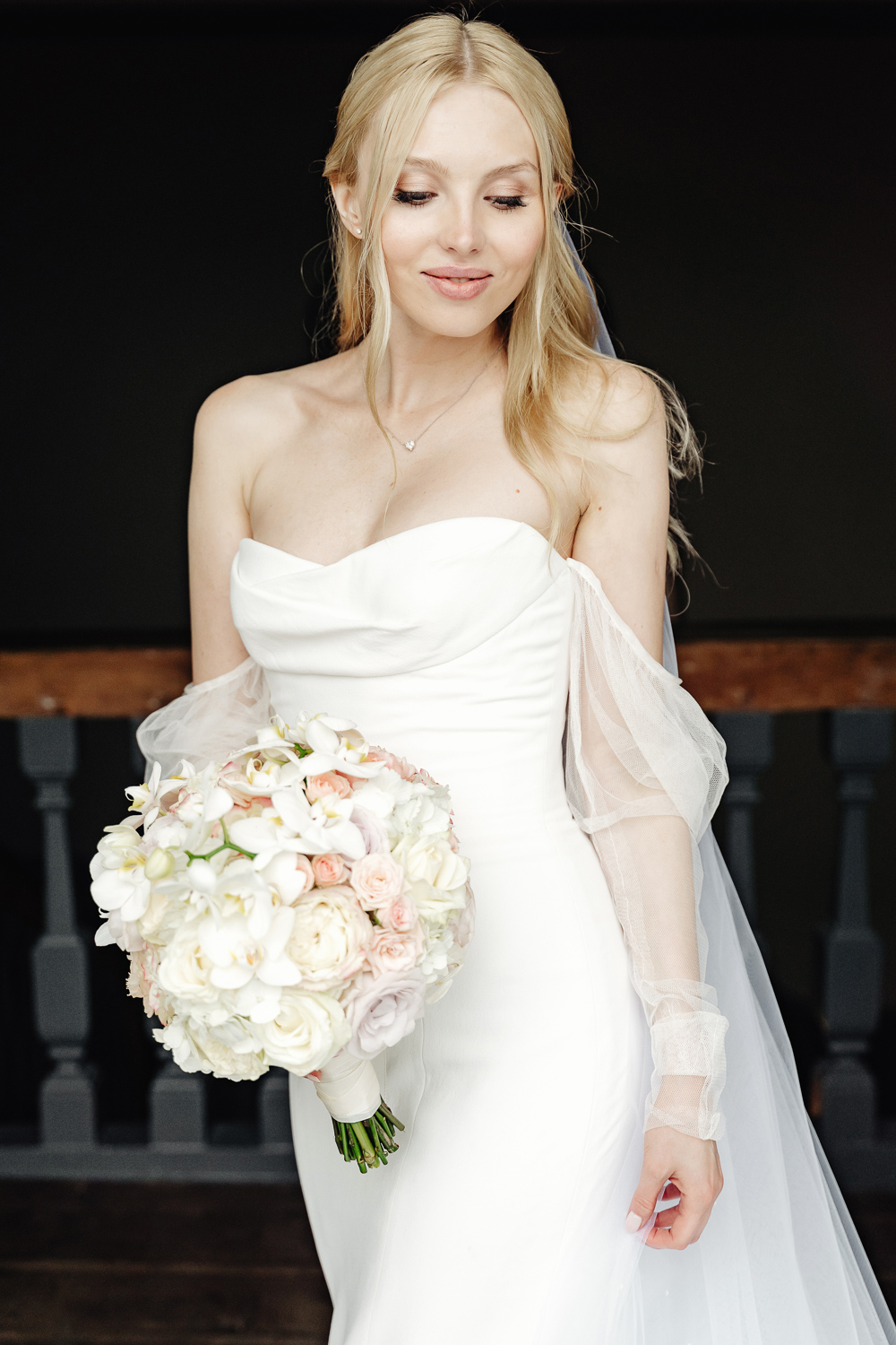 Beautiful blond bride stays in the balcony of castle Chateau Neercanne in Maastricht and slightly smiling and look down, with long veil and wedding dress Vera Wang by Mercury, holding flower bouquet